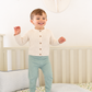 A smiling toddler stands on a patterned bed in a nursery, wearing a cream long-sleeve button-front cardigan and sage green pants.