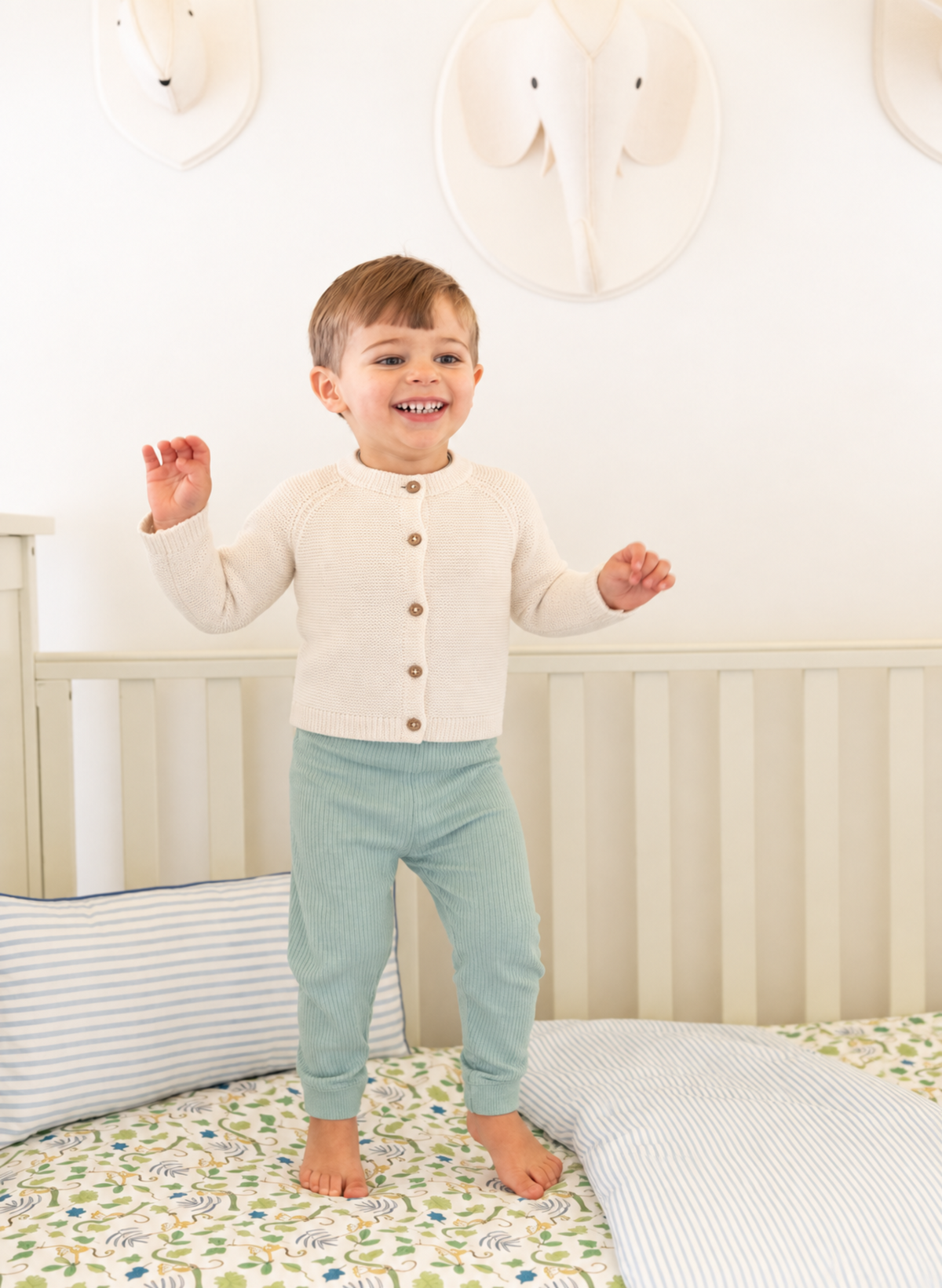 A smiling toddler stands on a patterned bed in a nursery, wearing a cream long-sleeve button-front cardigan and sage green pants.