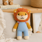 A knitted lion plush toy with an orange yarn mane, yellow arms and legs, and light blue overalls, sitting on a beige cushion in a styled indoor setting.