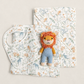 A baby gift set featuring a white muslin bib and a matching hooded wrap blanket with a whimsical safari animal print in muted blue and orange, styled with a small plush lion toy on a warm beige background.
