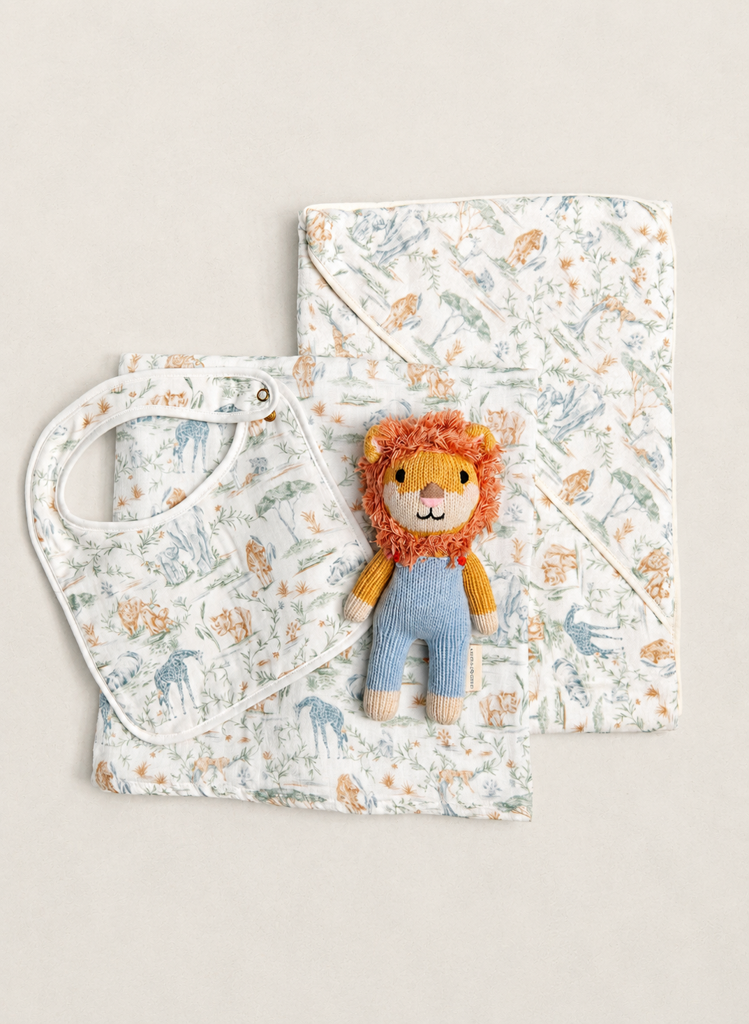 A baby gift set featuring a white muslin bib and a matching hooded wrap blanket with a whimsical safari animal print in muted blue and orange, styled with a small plush lion toy on a warm beige background.