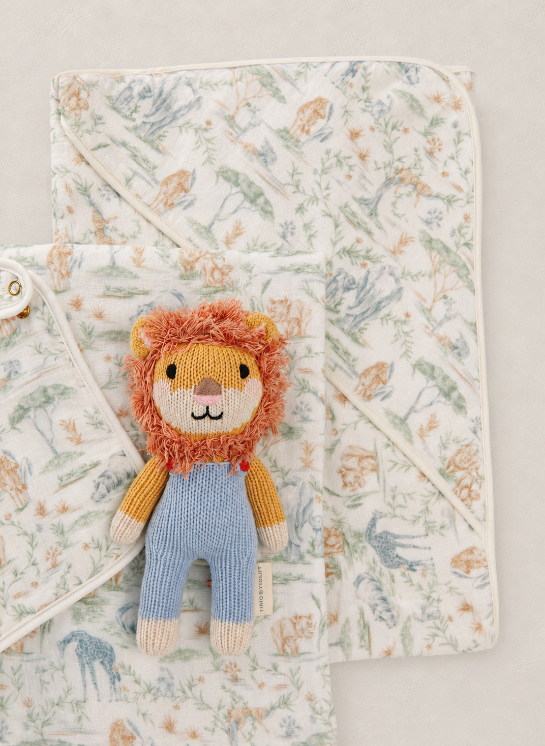 A soft baby blanket set with a white background and a delicate multicolor floral and animal print, styled with a small knitted lion plush toy.