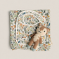 A folded baby gift set featuring a woodland-themed printed blanket, a matching bib, and a small plush monkey toy, arranged on a neutral beige background.