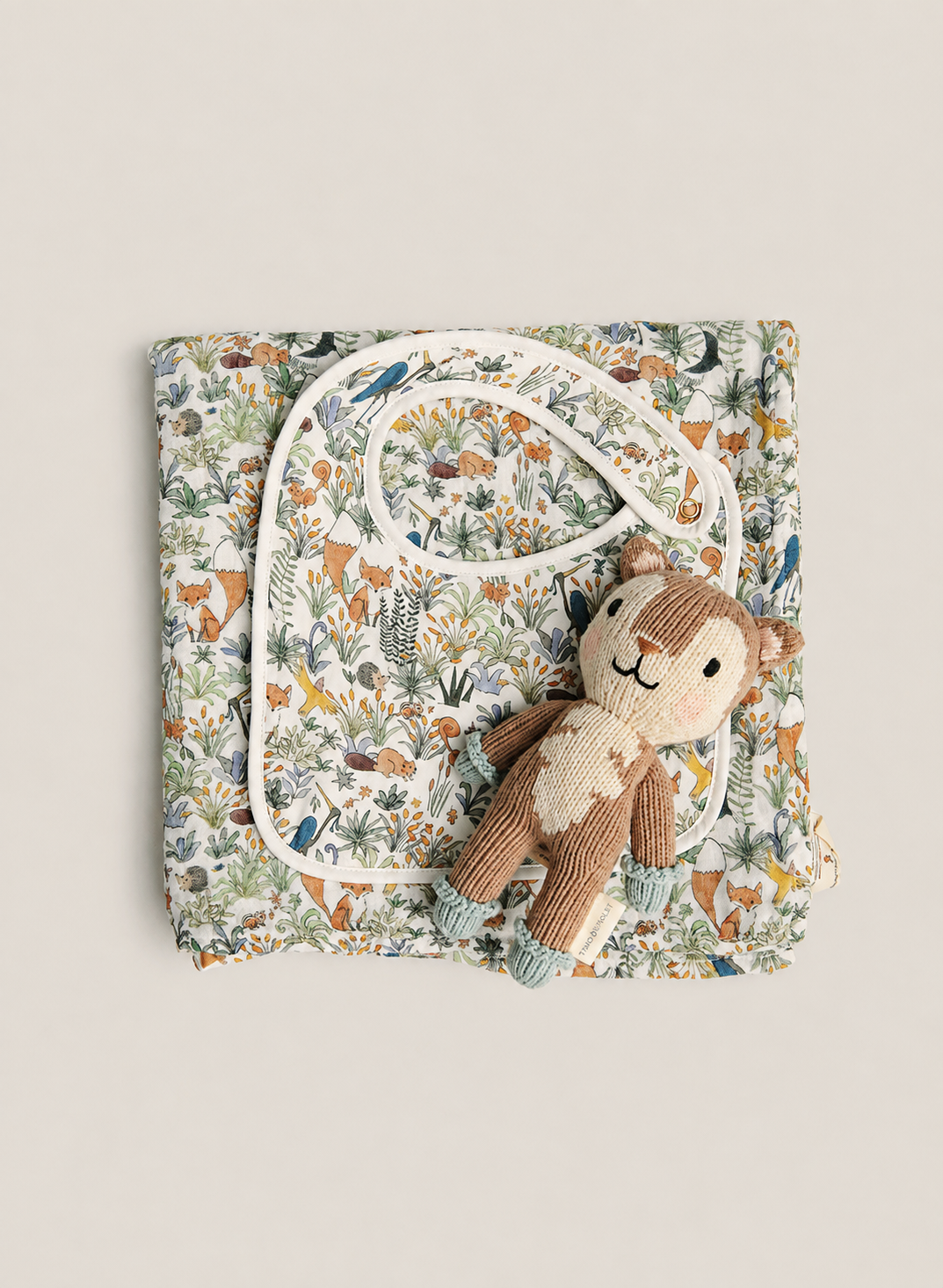 A folded baby gift set featuring a woodland-themed printed blanket, a matching bib, and a small plush monkey toy, arranged on a neutral beige background.