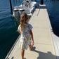 A young girl with blonde hair in a ponytail stands on a concrete dock next to the water, wearing a short-sleeve white dress with a colorful abstract print and beige sandals. A white boat is docked beside her.