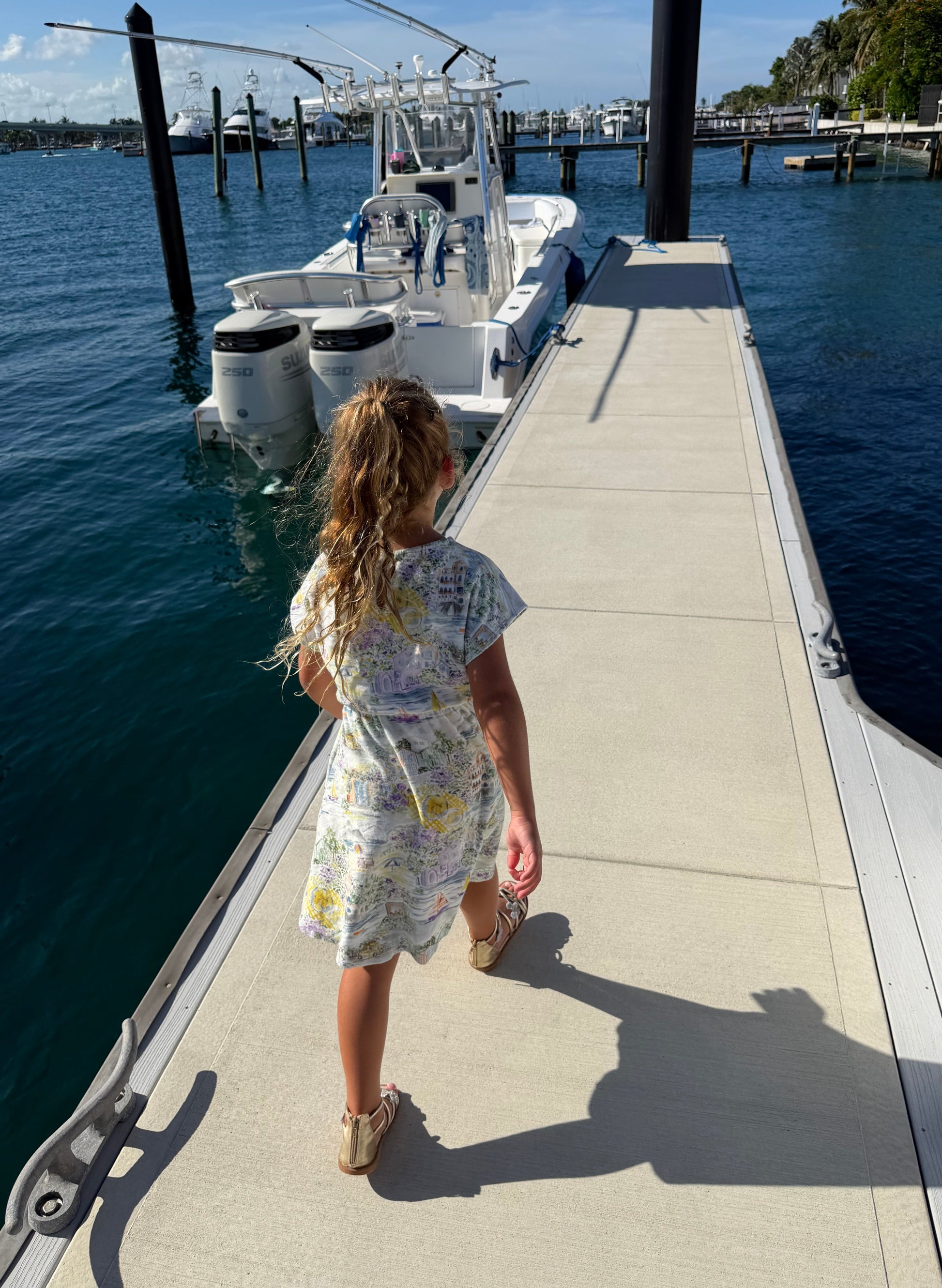 A young girl with blonde hair in a ponytail stands on a concrete dock next to the water, wearing a short-sleeve white dress with a colorful abstract print and beige sandals. A white boat is docked beside her.