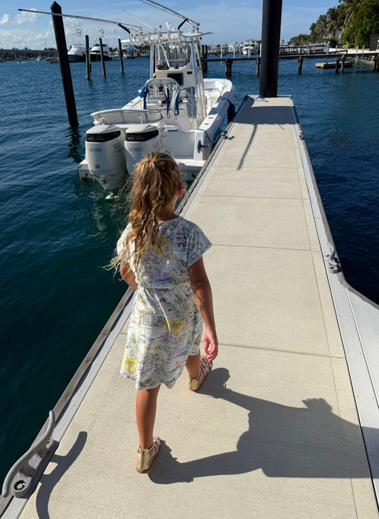 A young girl with blonde hair in a ponytail stands on a concrete dock next to the water, wearing a short-sleeve white dress with a colorful abstract print and beige sandals. A white boat is docked beside her.