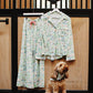 A matching two-piece pajama set with a light green and white floral print hangs on a rack against a wooden stable door, with a small brown dog wearing a matching bandana standing below.