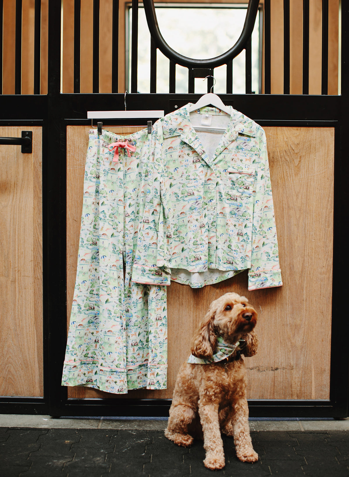 A matching two-piece pajama set with a light green and white floral print hangs on a rack against a wooden stable door, with a small brown dog wearing a matching bandana standing below.