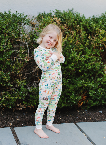 A young child wearing a long-sleeve matching pajama set with a colorful outdoor scene print, standing barefoot on a stone path in front of a green hedge.