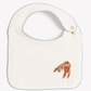 An off-white baby bib with a scalloped light blue border, featuring a small embroidered brown dog wearing a red bandana on the lower right.