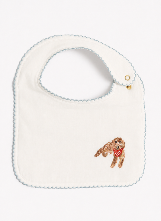 An off-white baby bib with a scalloped light blue border, featuring a small embroidered brown dog wearing a red bandana on the lower right.