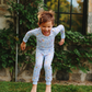 A young child wearing a light blue long-sleeve pajama set with a colorful speckled pattern, captured mid-jump on a grassy lawn in front of a stone wall and window.