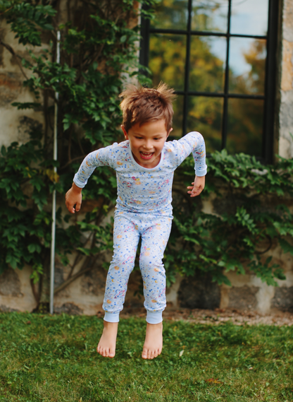A young child wearing a light blue long-sleeve pajama set with a colorful speckled pattern, captured mid-jump on a grassy lawn in front of a stone wall and window.