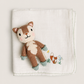 A folded white muslin baby blanket featuring a knitted brown and cream squirrel character and a small embroidered fox, displayed on a neutral beige background.