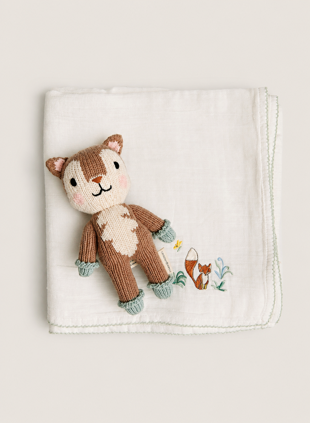 A folded white muslin baby blanket featuring a knitted brown and cream squirrel character and a small embroidered fox, displayed on a neutral beige background.