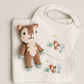 A baby gift set featuring a white muslin bib and matching burp cloth with embroidered woodland foxes, accompanied by a small knitted plush squirrel toy, arranged on a neutral beige background.