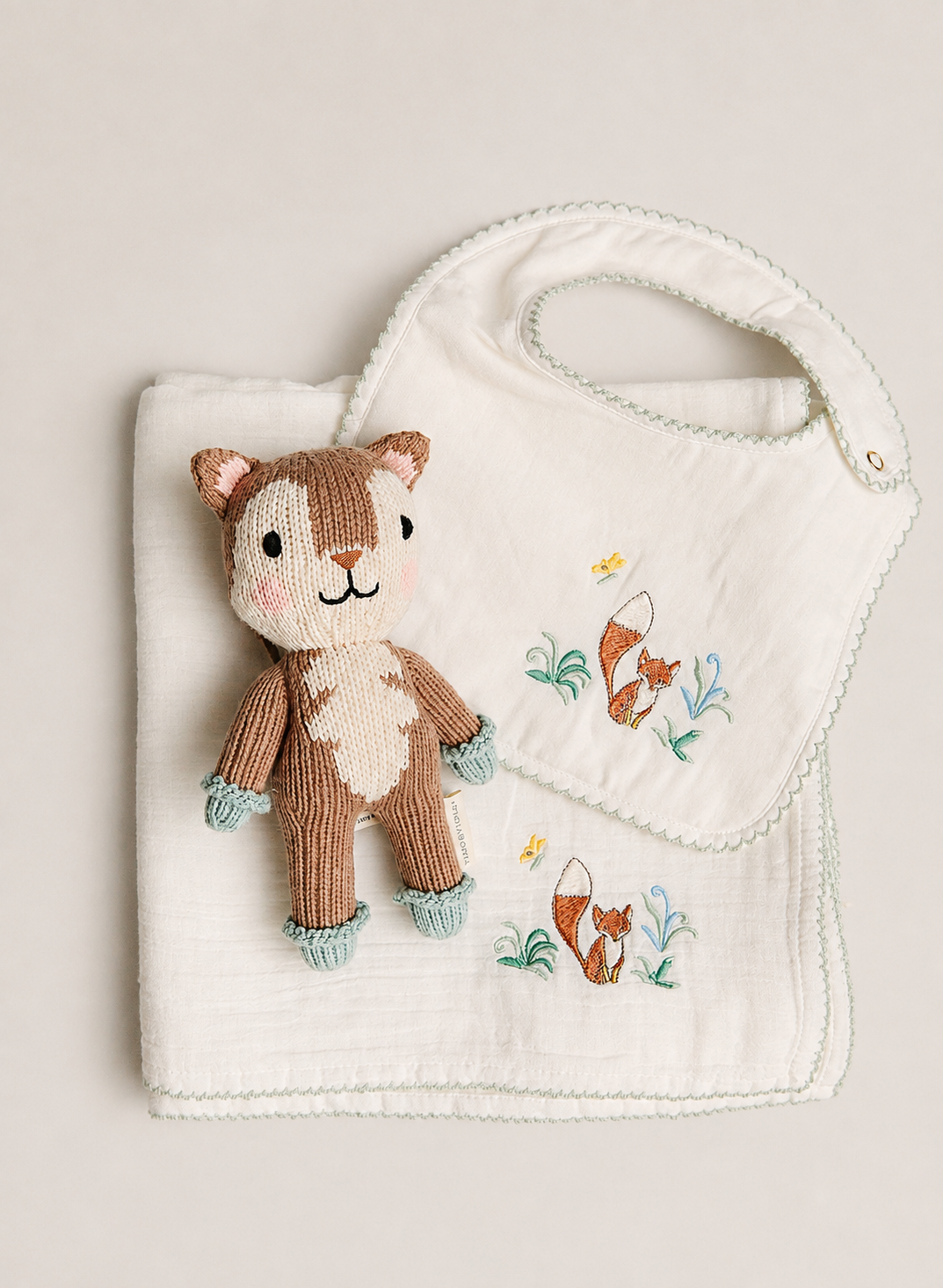 A baby gift set featuring a white muslin bib and matching burp cloth with embroidered woodland foxes, accompanied by a small knitted plush squirrel toy, arranged on a neutral beige background.