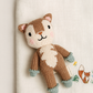 A knitted woodland animal toy with a brown and cream bear face and body, mint green paws and feet, displayed on a cream textured fabric backdrop with embroidered fox and butterfly details.