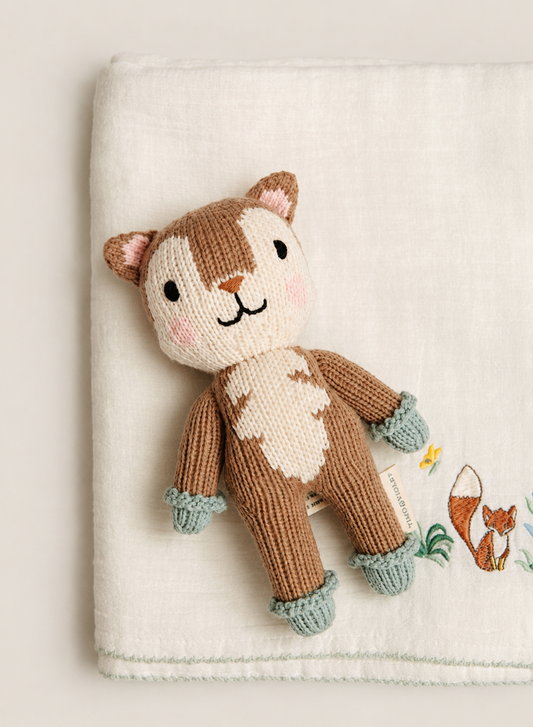 A knitted woodland animal toy with a brown and cream bear face and body, mint green paws and feet, displayed on a cream textured fabric backdrop with embroidered fox and butterfly details.