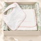 A baby gift set in a cream-colored box with the brand name 'Timo & Violet' printed on the front. The set includes a white muslin bib with pink pom-pom trim and embroidered text, a folded white blanket, and green botanical patterned tissue paper, all arranged on a light beige surface with soft leaf shadows.
