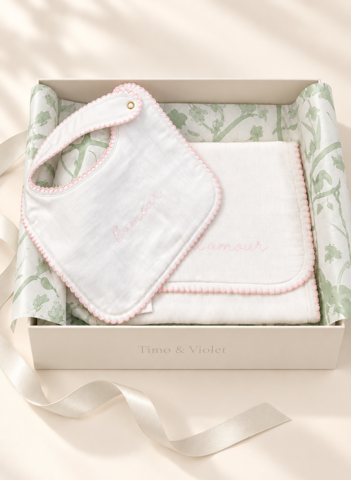 A baby gift set in a cream-colored box with the brand name 'Timo & Violet' printed on the front. The set includes a white muslin bib with pink pom-pom trim and embroidered text, a folded white blanket, and green botanical patterned tissue paper, all arranged on a light beige surface with soft leaf shadows.