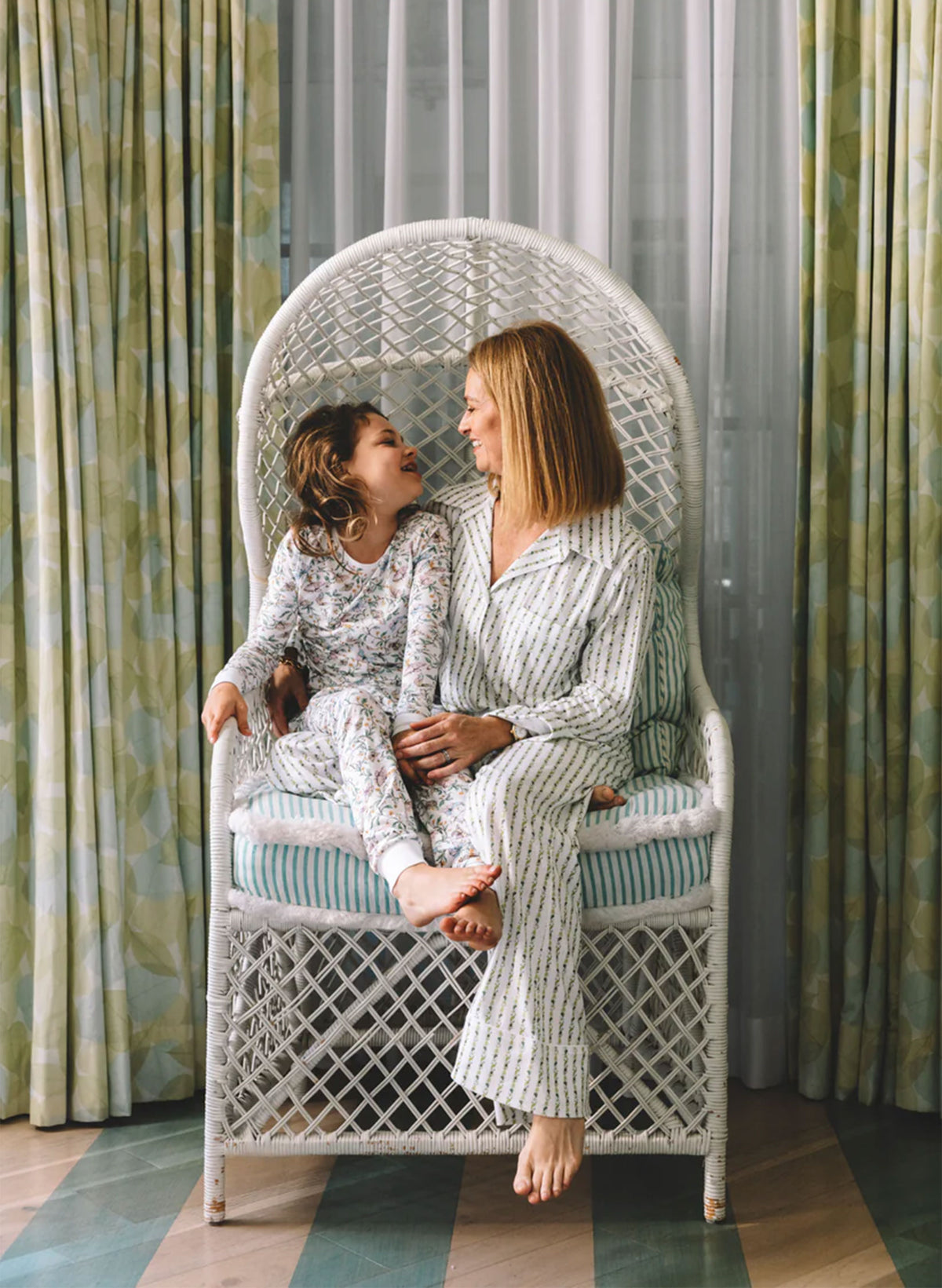Two women sitting on a wicker chair in a room with patterned curtains.
