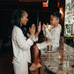 A woman and a young girl clapping happily at a bar. The woman wears a cream-colored pajama set with a subtle vertical stripe pattern, and the girl wears a matching floral print pajama set.