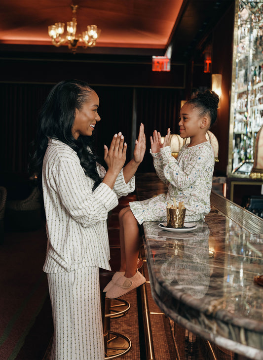 A woman and a young girl clapping happily at a bar. The woman wears a cream-colored pajama set with a subtle vertical stripe pattern, and the girl wears a matching floral print pajama set.
