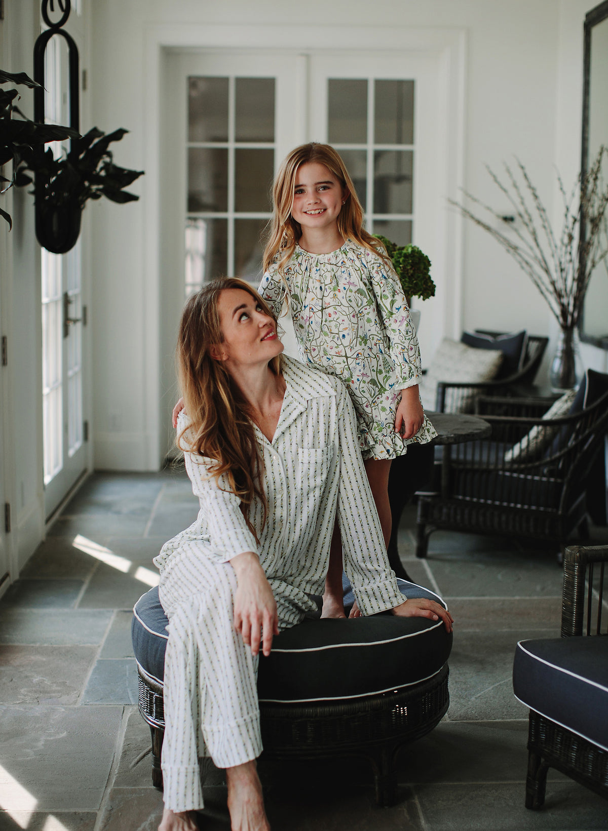 A woman and a young girl wearing matching floral pajama sets pose together in a bright, modern room with stone tile floors and large white French doors.