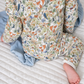 A baby wearing a long-sleeve jumpsuit with a detailed woodland animal print featuring foxes, birds, and foliage on an off-white background, sitting on a white quilted mattress with a light blue blanket.