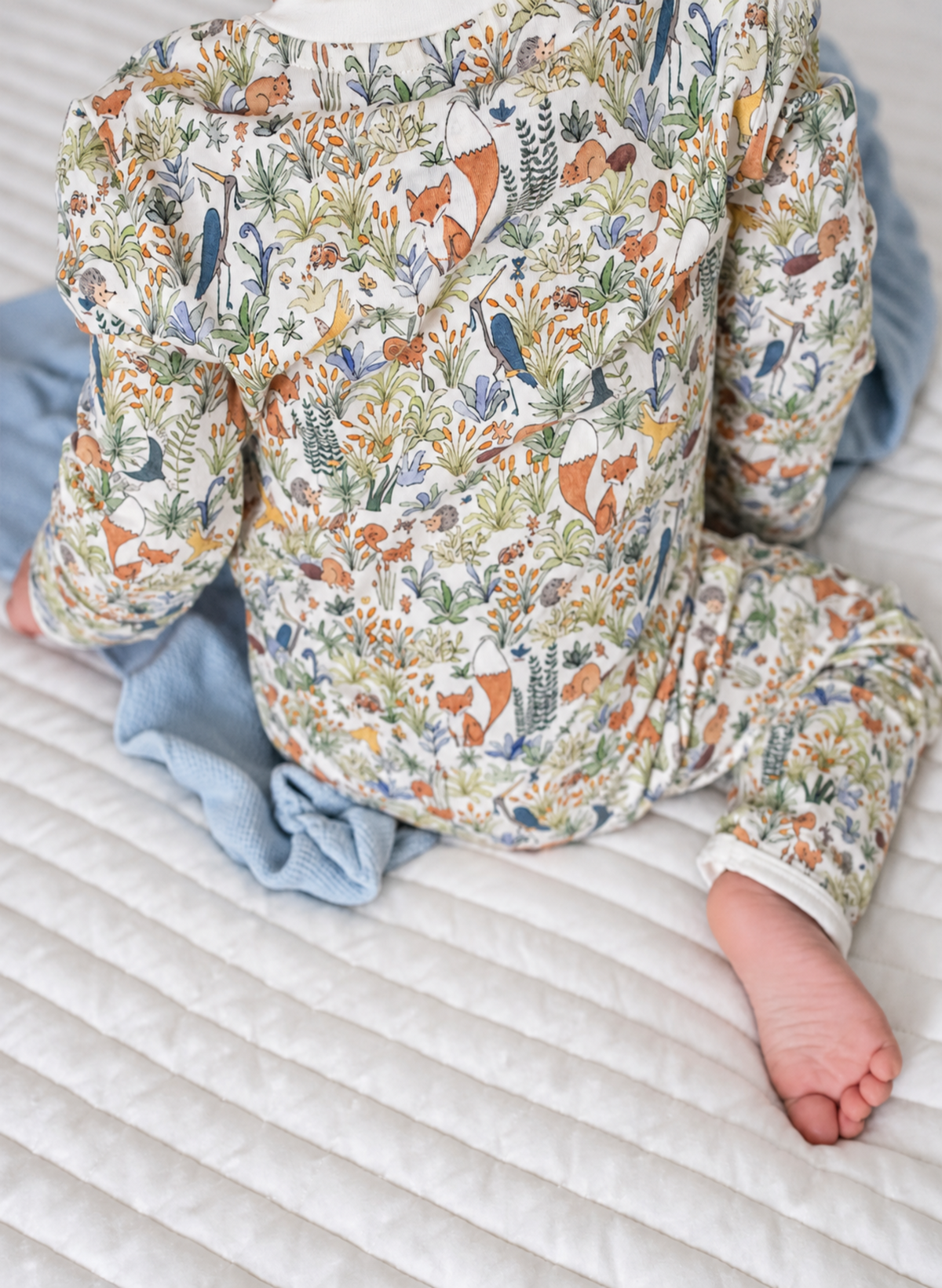 A baby wearing a long-sleeve jumpsuit with a detailed woodland animal print featuring foxes, birds, and foliage on an off-white background, sitting on a white quilted mattress with a light blue blanket.