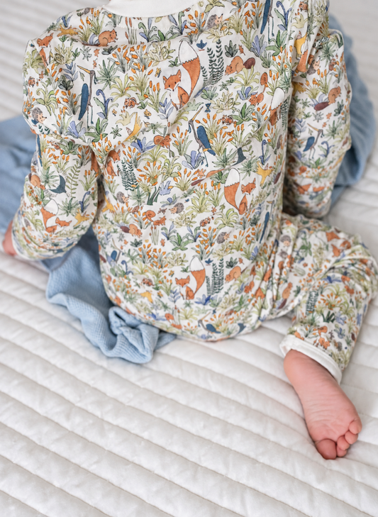 A baby wearing a long-sleeve jumpsuit with a detailed woodland animal print featuring foxes, birds, and foliage on an off-white background, sitting on a white quilted mattress with a light blue blanket.