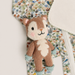 A close-up of a knitted animal plush toy with a brown and cream body, placed over a patterned garment featuring a woodland print with birds, foxes, and foliage.