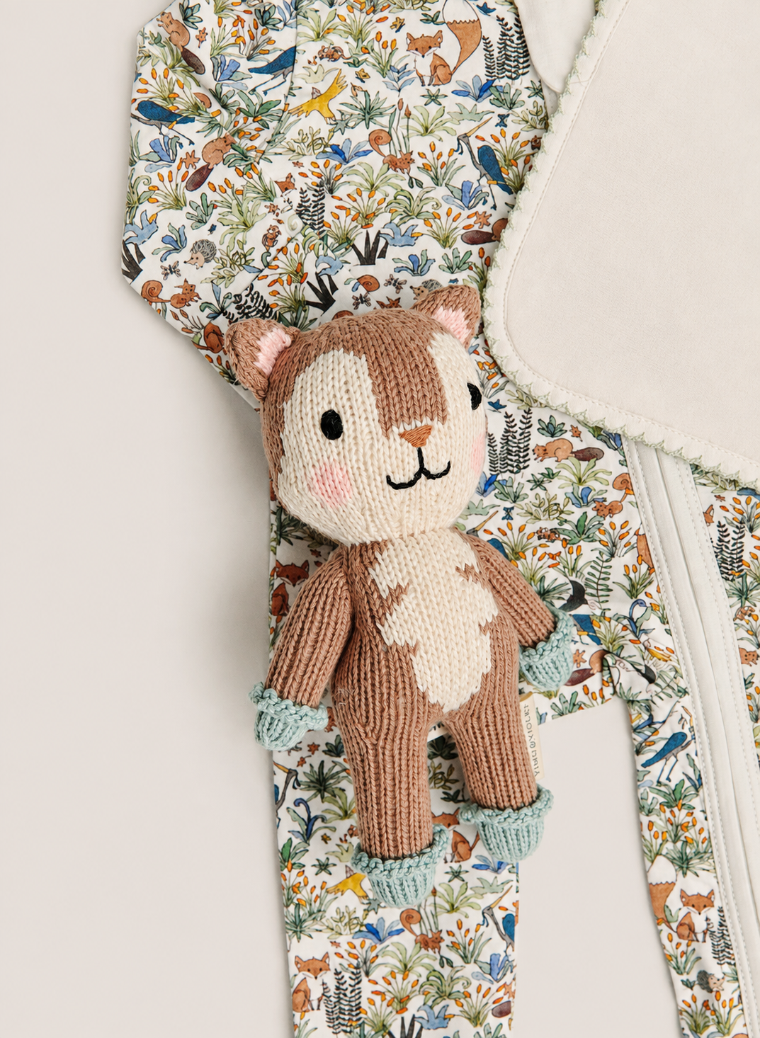A close-up of a knitted animal plush toy with a brown and cream body, placed over a patterned garment featuring a woodland print with birds, foxes, and foliage.