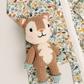 A close-up of a knitted deer plush toy placed on top of a floral and woodland print zippered garment. The toy is brown and cream with pink cheeks and light blue booties, while the background fabric has a multicolor nature print with birds, foxes, and foliage.