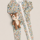 A baby zip-up one-piece romper with a dense woodland animal print, featuring a white zipper down the front, laid flat on a light beige background with a small plush squirrel toy resting on the chest.
