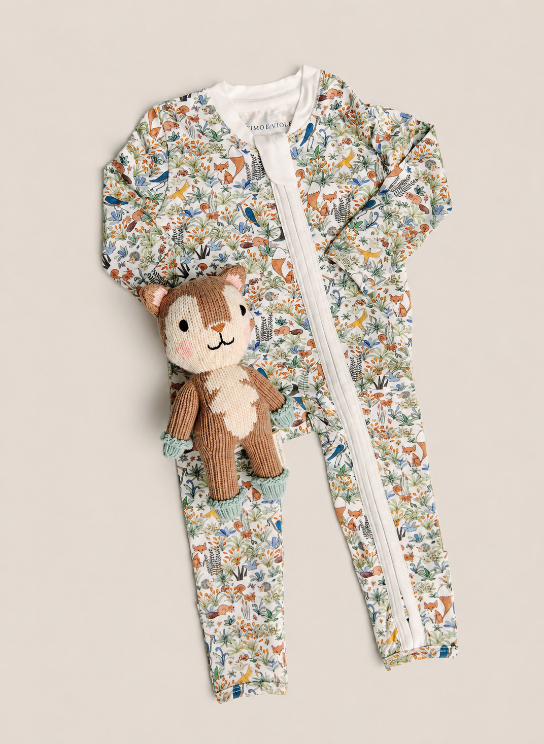 A baby zip-up one-piece romper with a dense woodland animal print, featuring a white zipper down the front, laid flat on a light beige background with a small plush squirrel toy resting on the chest.