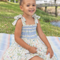 A young girl with two buns and blue bows sits on a striped blanket in a park, wearing a smocked sleeveless dress with a blue and white patterned bodice and a white floral skirt.