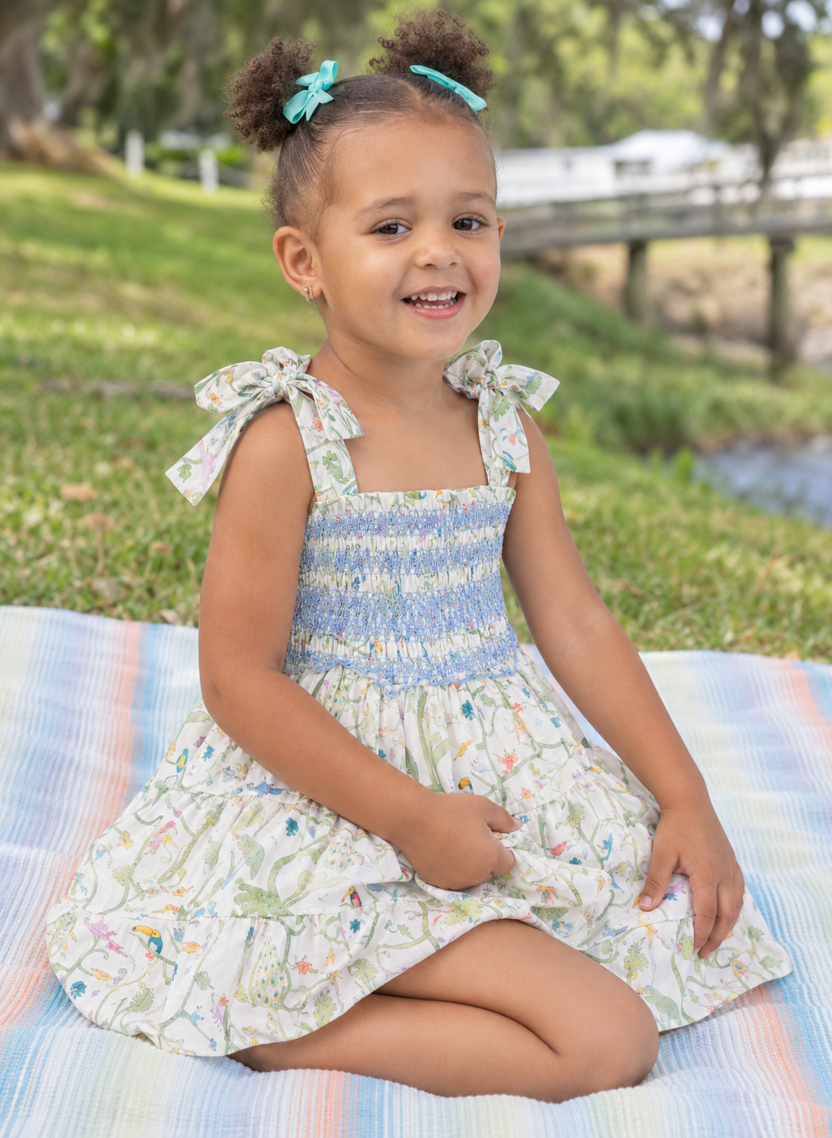 A young girl with two buns and blue bows sits on a striped blanket in a park, wearing a smocked sleeveless dress with a blue and white patterned bodice and a white floral skirt.