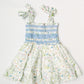 A sleeveless children's dress with a smocked bodice, ruffled shoulder straps, and a tiered skirt, laid flat on a light gray background. The dress features a white base with a pastel floral and vine print in green, pink, blue, and yellow, with a light blue smocked waist section.