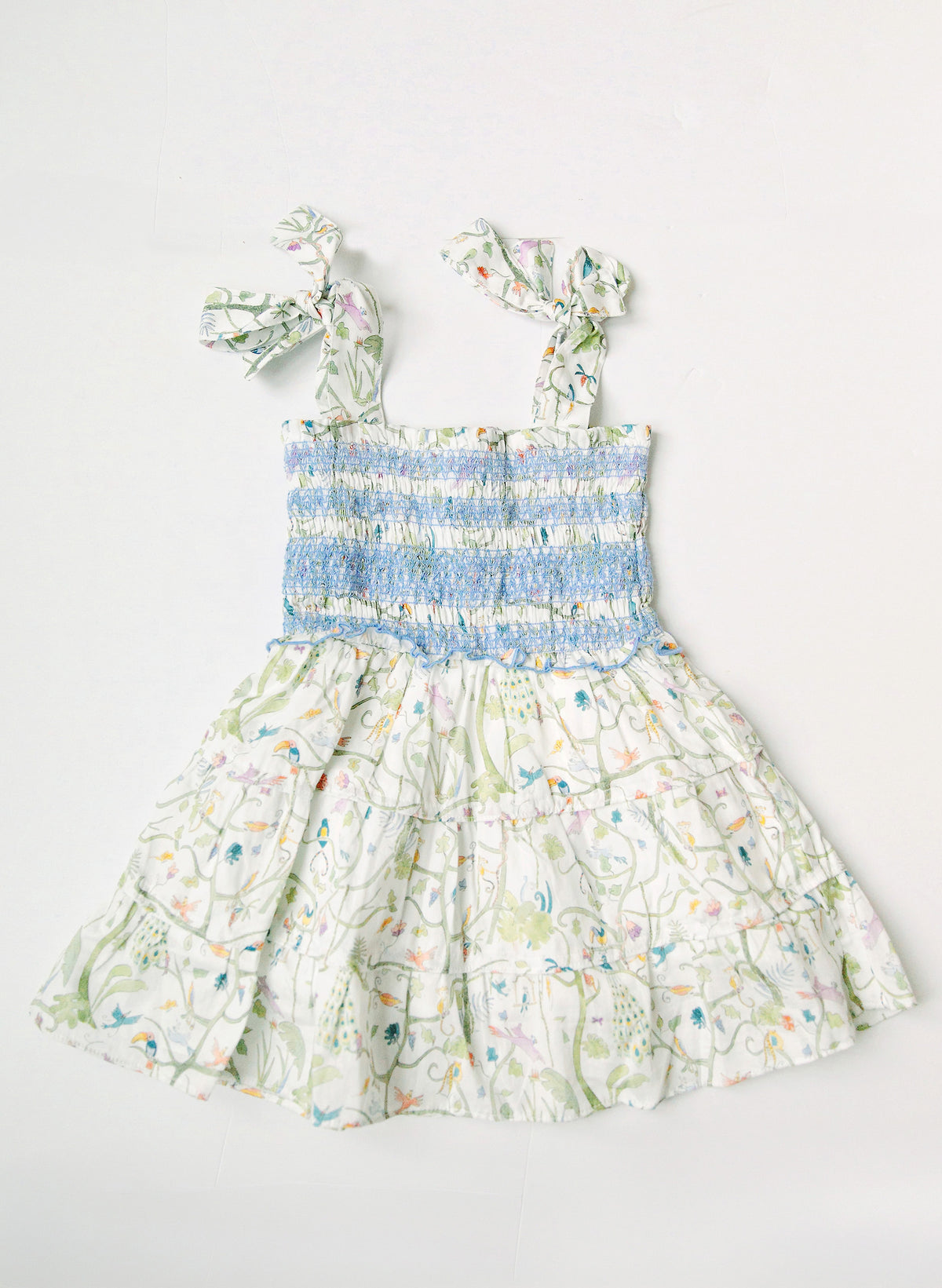 A sleeveless children's dress with a smocked bodice, ruffled shoulder straps, and a tiered skirt, laid flat on a light gray background. The dress features a white base with a pastel floral and vine print in green, pink, blue, and yellow, with a light blue smocked waist section.
