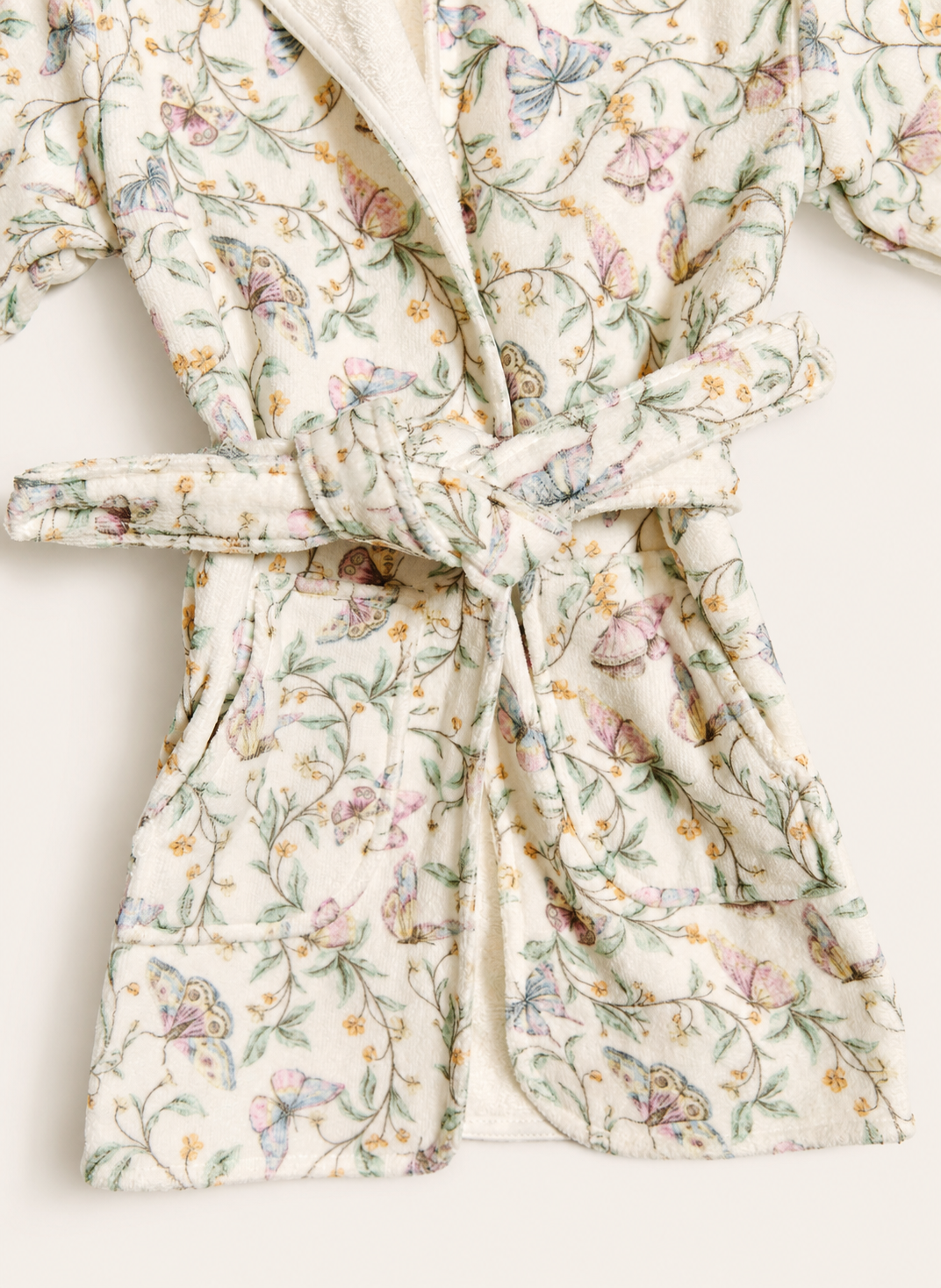 A close-up of a quilted bathrobe with a tie belt, featuring a delicate floral and butterfly pattern in soft pastel colors on an off-white background.