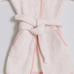 A close-up of a light pink children's robe with a matching tie belt, featuring a subtle embossed circular pattern and pink piping along the edges.