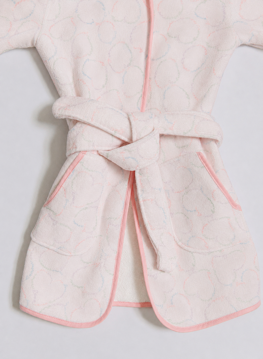 A close-up of a light pink children's robe with a matching tie belt, featuring a subtle embossed circular pattern and pink piping along the edges.