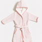 A light pink hooded bathrobe with a matching tie belt, featuring a subtle tonal pattern, pink piping, and a white inner lining, displayed flat against a light gray background.