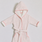 A light pink hooded bathrobe with a subtle tonal pattern, featuring pink piping, a matching tie belt, and white plush lining inside the hood and cuffs, laid flat against a light gray background.