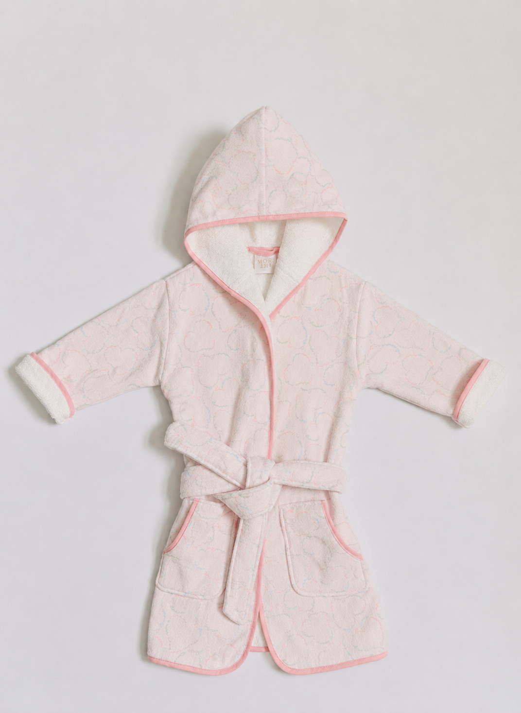 A light pink hooded bathrobe with a subtle tonal pattern, featuring pink piping, a matching tie belt, and white plush lining inside the hood and cuffs, laid flat against a light gray background.