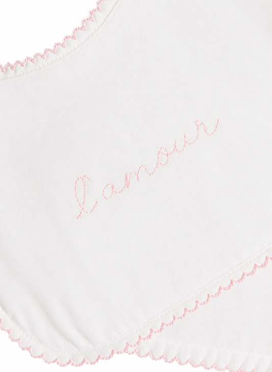 Close-up of a white fabric item with pink scalloped trim and the word 'amour' embroidered in pink script across the center.
