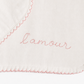 Close-up of a white fabric item with pink scalloped stitching and the word 'amour' embroidered in pink script across the center.